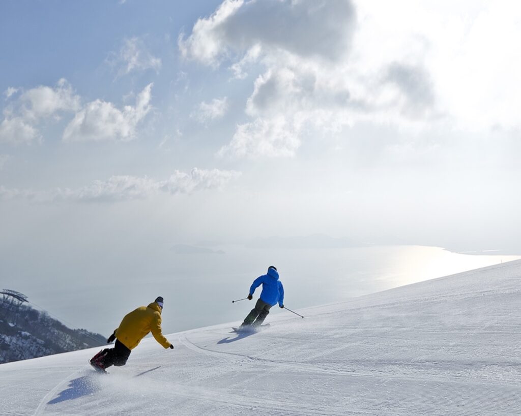 Two skiers carving down a groomed run at Biwako Valley, with Lake Biwa shining below in Shiga