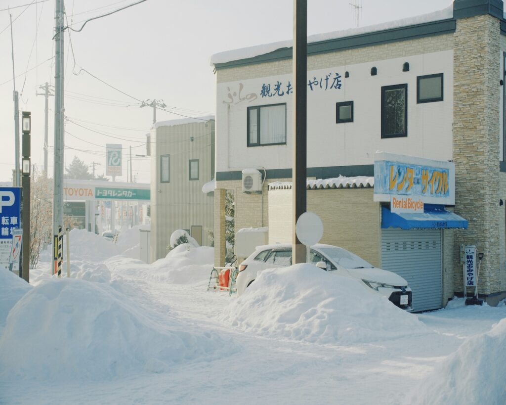 Snow piled high along a street in Furano, Hokkaido, with a souvenir shop and a car buried in snow