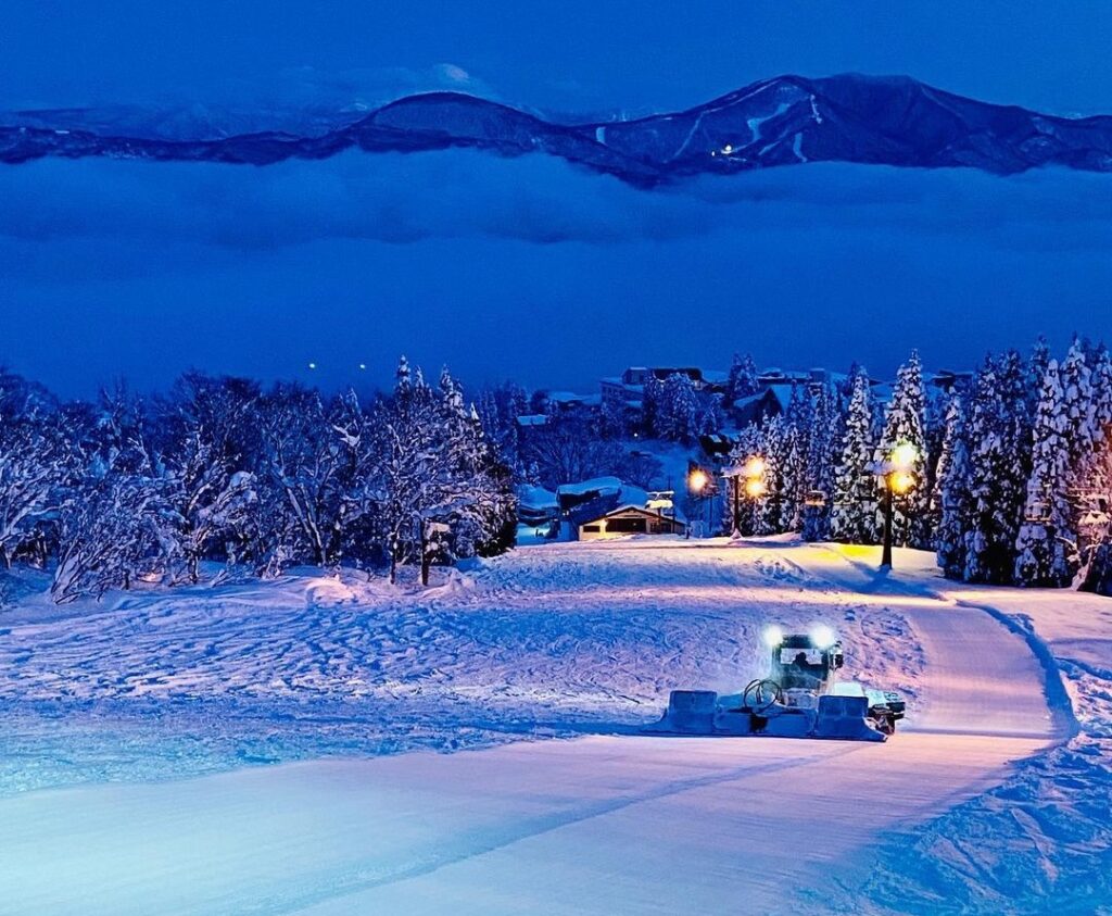 Snow groomer on a lit ski slope at dusk in Myoko Kogen, Niigata, with mountains above a sea of clouds