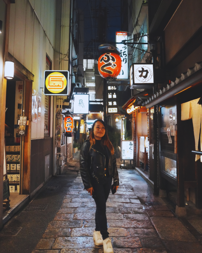 Flip Japan photo of a woman standing in Namba at night lit up by neon signs for Osaka vs Tokyo