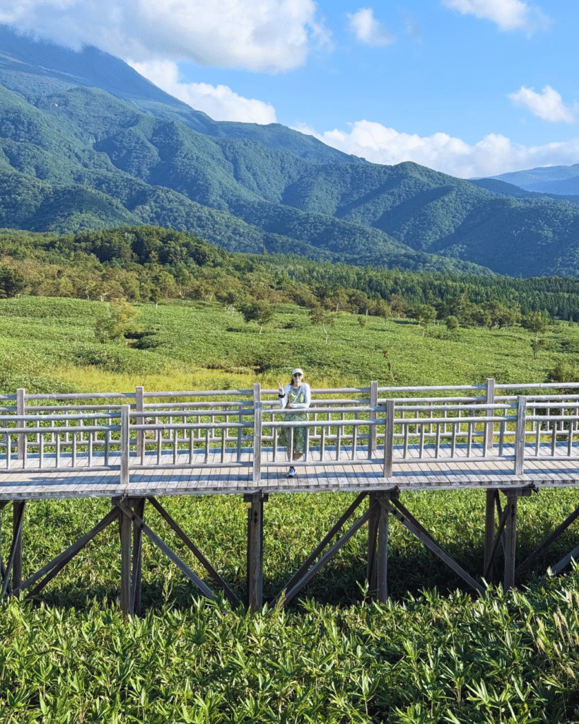 Photo of a woman standing on a raised path in Shiretoko with tall grass below and mountains behind, one of the most scenic places in Japan.