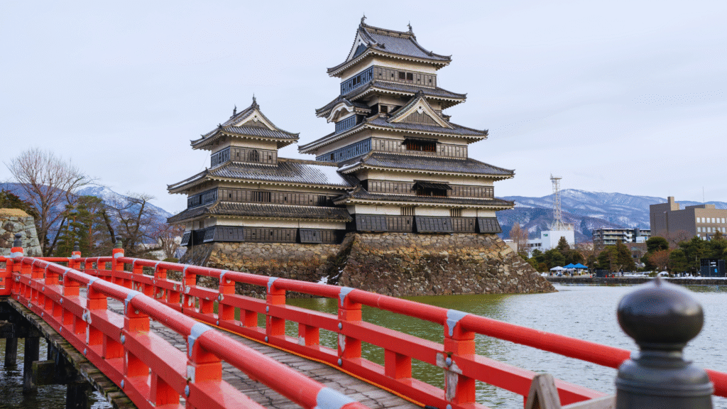 Photo of the ancient Japanese castle of Matsumoto, with a red bridge in the foreground, a moat, and mountains behind