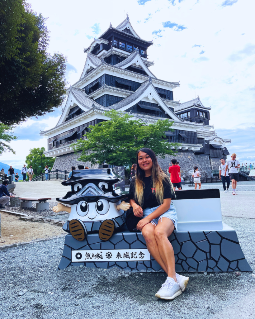 Flip Japan photo of Venese sitting in front of Kumamoto castleon a bench with a statue of the Kumamoto castle mascot