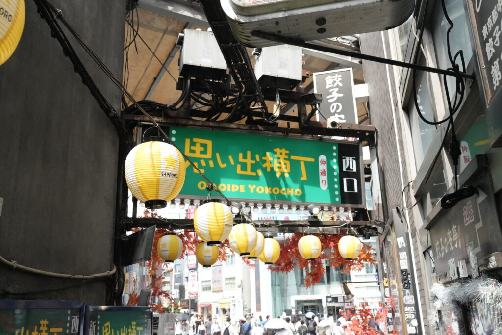 Flip Japan photo of Omoide Yokocho in Shinjuku with red maple leaf autumn decorations and yellow lanterns overhead.