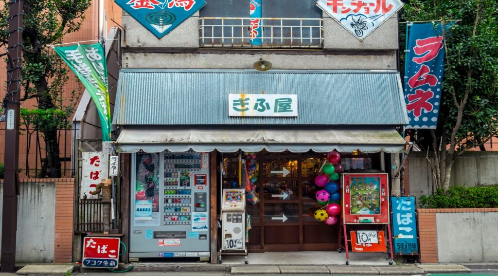 Showa retro storefront with large colorful signs, block Japanese text, vending machines and balloons outside.