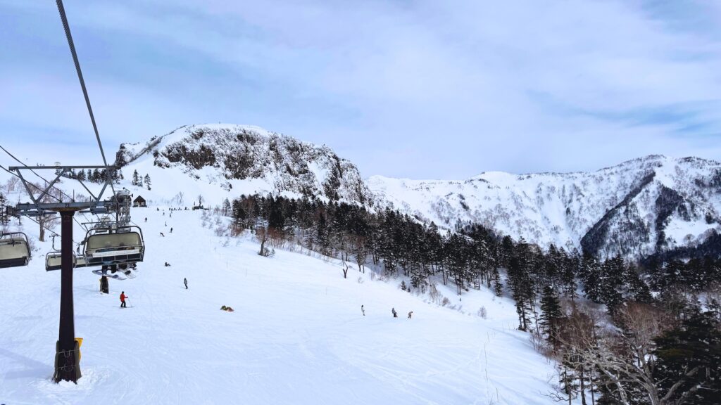 Chairlift above a wide groomed run with skiers, below a rocky snow-covered peak at Kawaba Ski Resort near Tokyo
