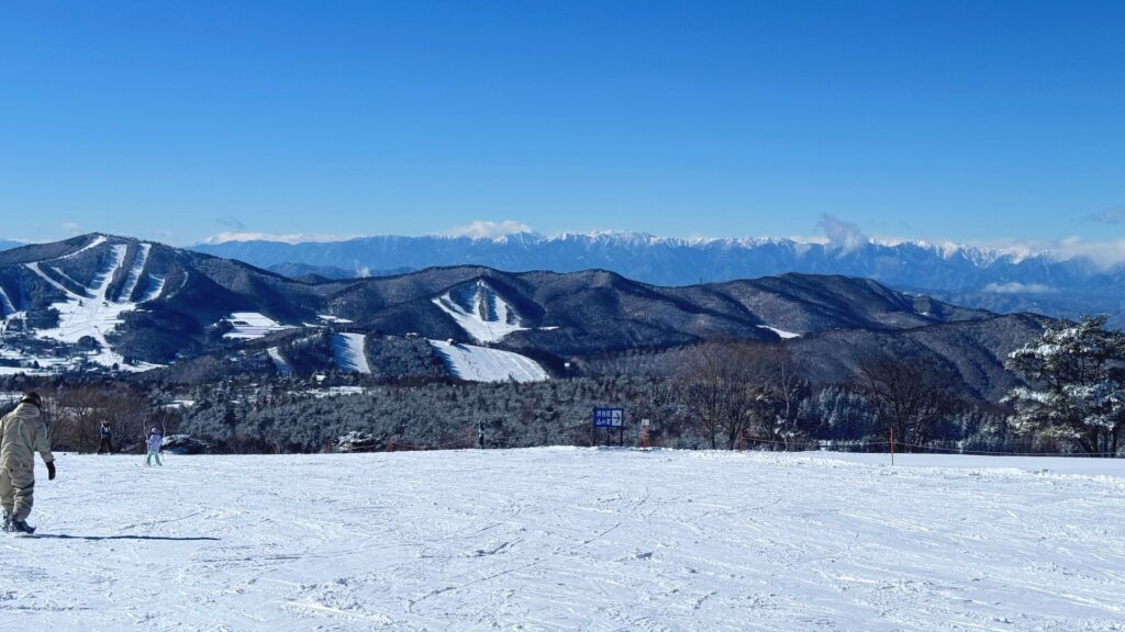 Sunny groomed slope with ski runs on nearby hills and a snow-capped mountain range on the horizon at Sugadaira Kogen Snow Resort, Nagano