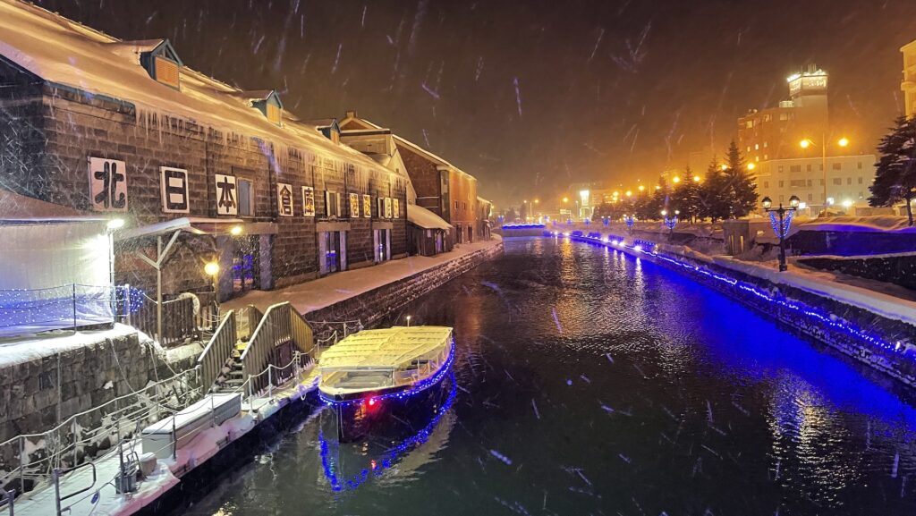 Otaru Canal at night in winter, with snow falling over the old stone warehouses and a canal boat lit in blue